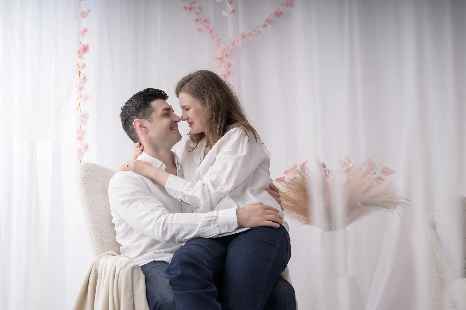 sedinta foto cuplu Cuplu indragostit zambind si privindu-se pe fotoliu alb, decor studio romantic, sedinta foto cuplu