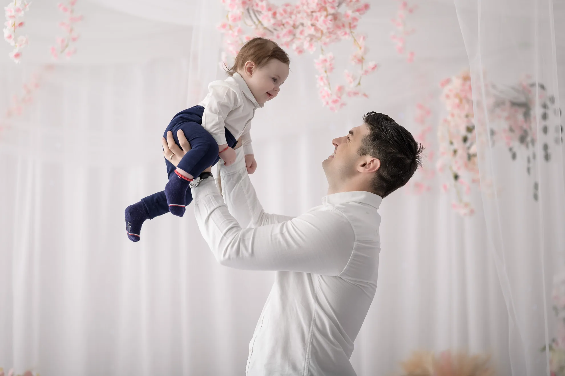 Tata ridicand bebelusul spre cer intr-un decor cu flori roz, sedinta foto familie bucuresti plina de emotie